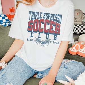 Op de afbeelding: Wit T-shirt met een vintage-stijl grafische afbeelding met de tekst "Triple Espresso Soccer Club" met een voetbal en vleugels. De tekst "Estd. 2024" staat onder de afbeelding.