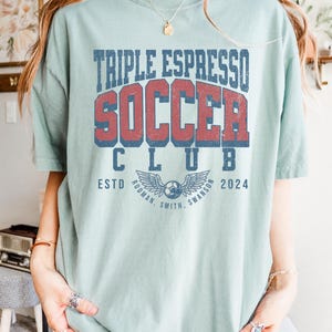 Op de afbeelding: Een lichtblauw T-shirt met de tekst "Triple Espresso Soccer Club Est. 2024" in een vintage lettertype. De tekst is in rood en blauw met een afbeelding van een voetbal en vleugels eronder.