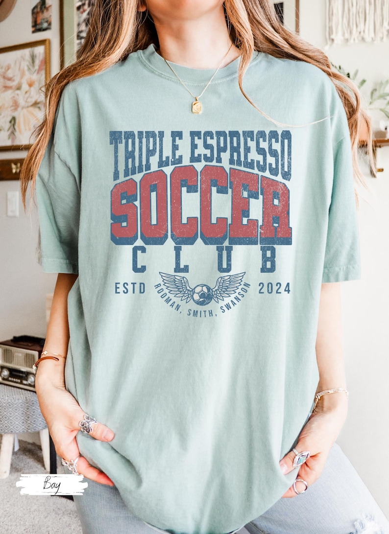 Op de afbeelding: Een lichtblauw T-shirt met de tekst "Triple Espresso Soccer Club Est. 2024" in een vintage lettertype. De tekst is in rood en blauw met een afbeelding van een voetbal en vleugels eronder.