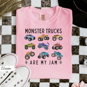 May include: A pale pink t-shirt featuring the phrase "MONSTER TRUCKS ARE MY JAM" and illustrations of monster trucks in various colours. The shirt is made of a soft material and is a comfortable fit.