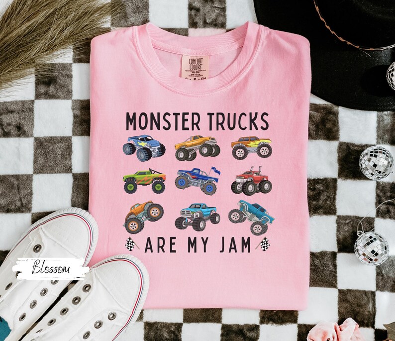 May include: A pale pink t-shirt featuring the phrase "MONSTER TRUCKS ARE MY JAM" and illustrations of monster trucks in various colours. The shirt is made of a soft material and is a comfortable fit.