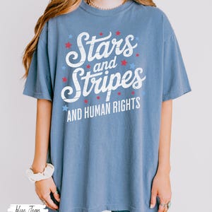 Puede incluir: Camiseta azul con el texto "Stars and Stripes AND HUMAN RIGHTS" en blanco, rojo y azul. La camiseta tiene un corte holgado y mangas cortas. El dise&ntilde;o incluye estrellas.