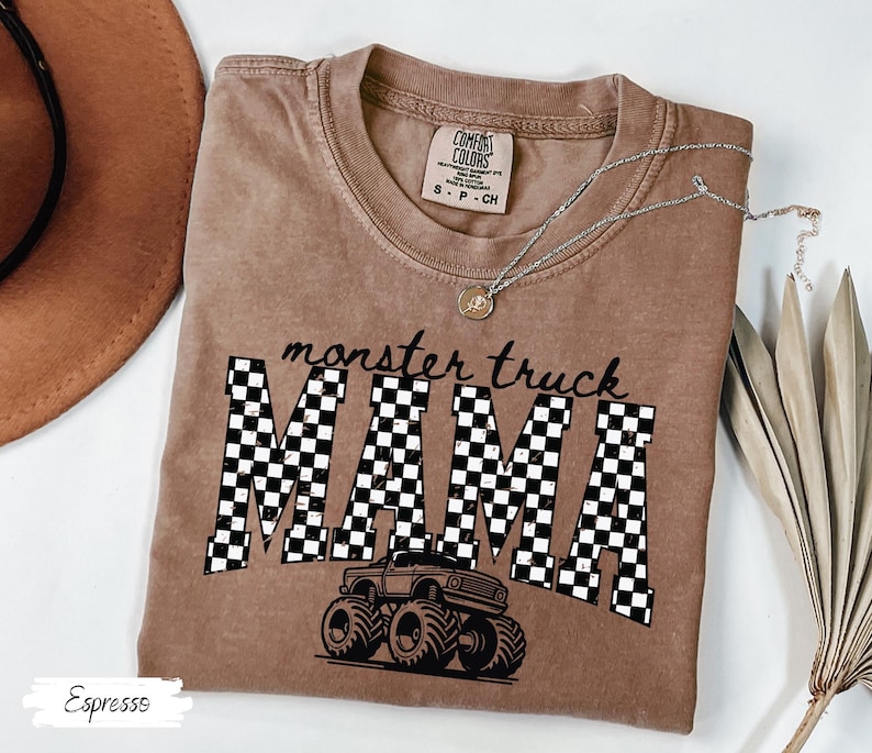 Op de afbeelding: Espresso-kleurig T-shirt met de tekst "monster truck MAMA" in zwart schrift en geblokte letters. Een monster truck-afbeelding staat onder de tekst. Een zilveren ketting met een hanger ligt op het shirt.