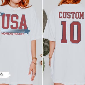 May include: White t-shirt with "USA WOMENS HOCKEY" in red and blue lettering. The back of the shirt features the word "CUSTOM" above the number "10" in red.