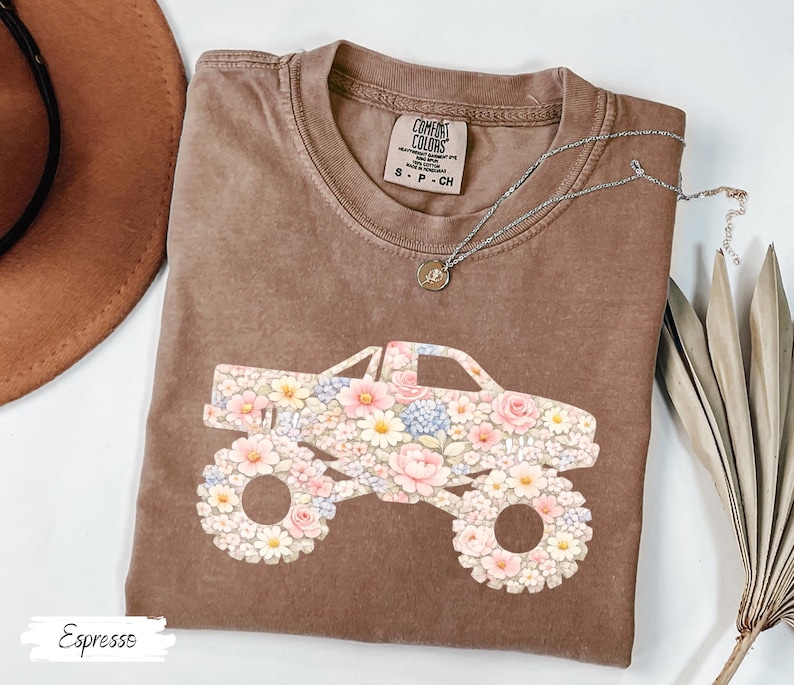 Op de afbeelding: Espresso-kleurig t-shirt met een bloemen monster truck ontwerp. De truck is versierd met roze, witte en blauwe bloemen. Het shirt heeft een ronde hals en korte mouwen. Een zilveren ketting met een kleine hanger ligt op het shirt.