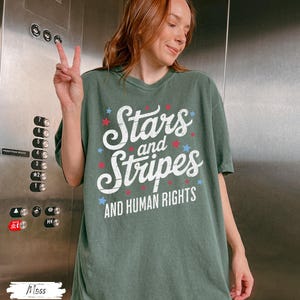 Puede incluir: Una camiseta verde con las palabras "Stars and Stripes AND HUMAN RIGHTS" en blanco, rojo y azul. La camiseta tiene un corte holgado y la lleva una persona.