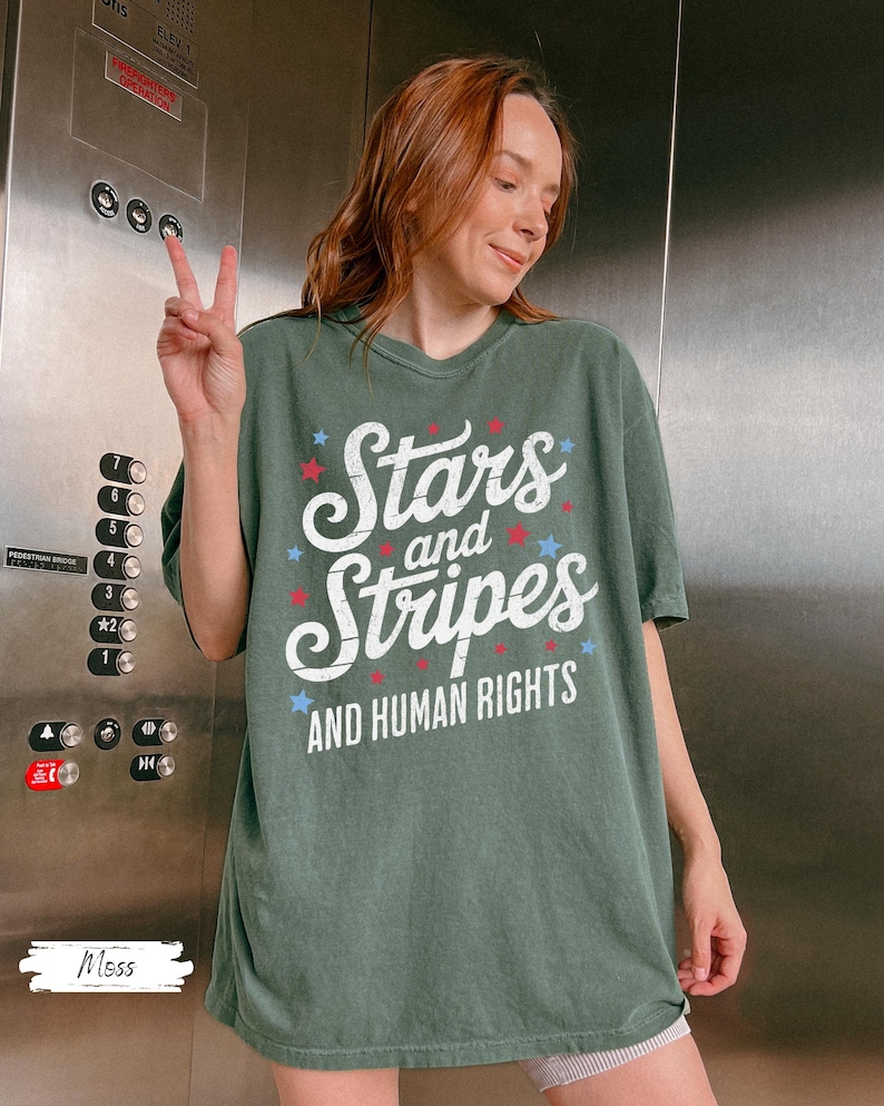 Puede incluir: Una camiseta verde con las palabras "Stars and Stripes AND HUMAN RIGHTS" en blanco, rojo y azul. La camiseta tiene un corte holgado y la lleva una persona.