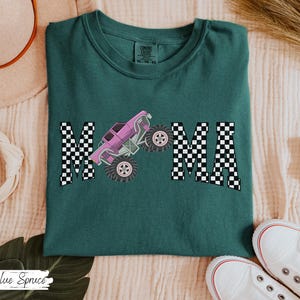 May include: Teal Comfort Colors t-shirt with the word "MAMA" in a checkered pattern. A pink monster truck graphic is centered between the letters. White sneakers are visible at the bottom right. The shirt is a size XL.