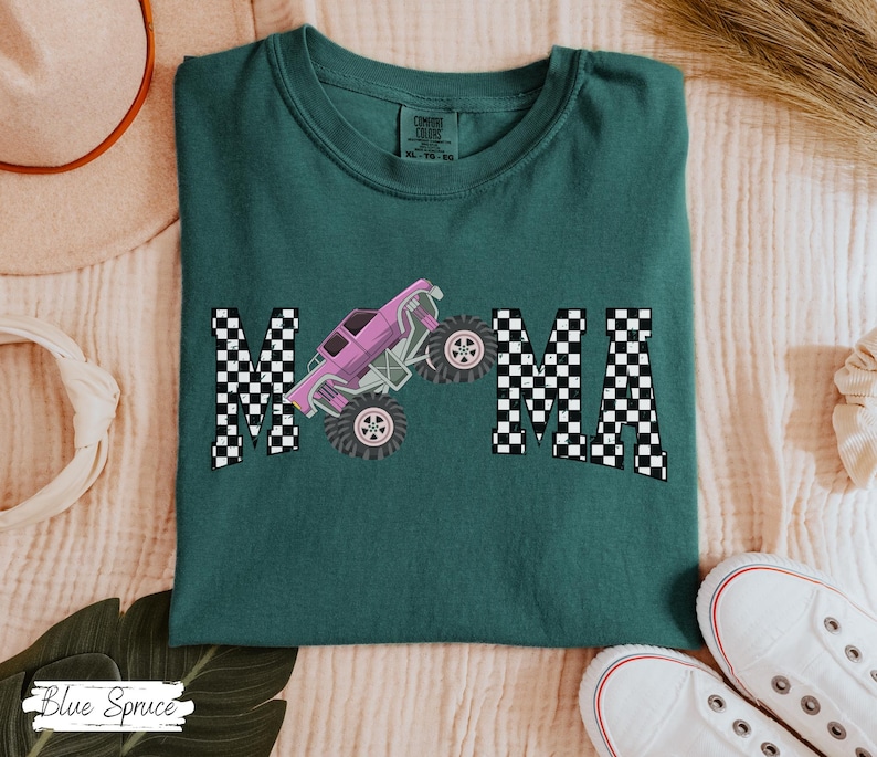 May include: Teal Comfort Colors t-shirt with the word "MAMA" in a checkered pattern. A pink monster truck graphic is centered between the letters. White sneakers are visible at the bottom right. The shirt is a size XL.