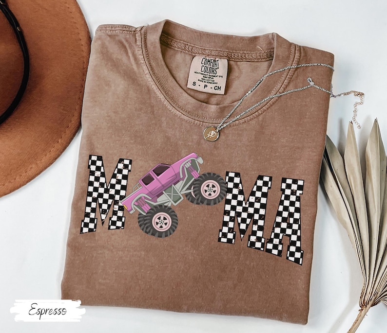 May include: Espresso-colored t-shirt with the word "MAMA" in a checkered pattern, featuring a pink monster truck graphic. The shirt has a crew neck and short sleeves. A silver necklace with a pendant is draped on the shirt.