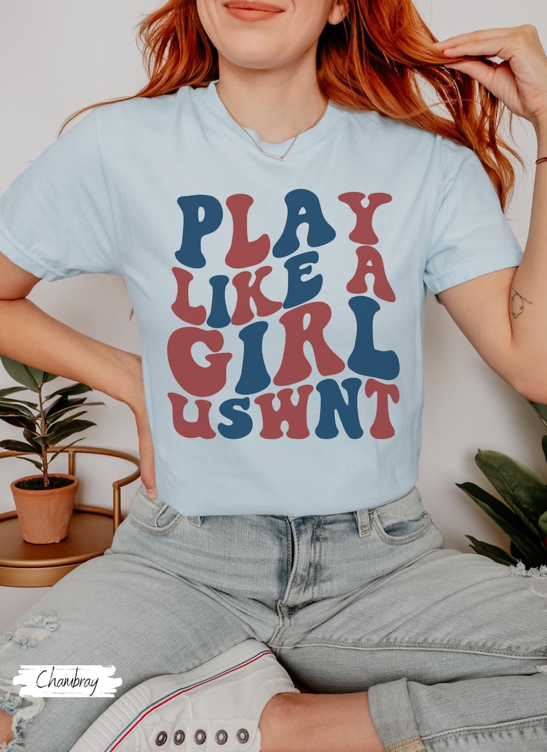 May include: A light blue t-shirt with the text "PLAY LIKE A GIRL USHWN" in a retro font. The text is in red and blue.