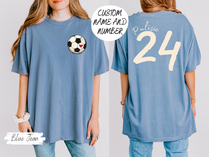 Op de afbeelding: Een blauw spijkerbroekkleurig T-shirt met een voetbalafbeelding en een hart. De achterkant van het shirt toont de naam "Pulisic" en het nummer "24" in cr&egrave;me. Het shirt heeft een comfortabele, oversized pasvorm.