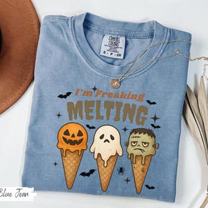 May include: Blue jean colored t-shirt with the text "I'm Freaking Melting" and three ice cream cones designed as a jack-o'-lantern, ghost, and Frankenstein's monster. The shirt has a crew neck and short sleeves. A gold necklace is draped across the shirt.