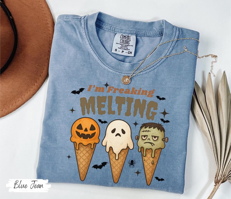 May include: Blue jean colored t-shirt with the text "I'm Freaking Melting" and three ice cream cones designed as a jack-o'-lantern, ghost, and Frankenstein's monster. The shirt has a crew neck and short sleeves. A gold necklace is draped across the shirt.