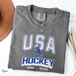 May include: A gray Comfort Colors t-shirt with a USA Hockey graphic in white and blue. The design includes a hockey player illustration and the years 1980 and 2026. A gold necklace is draped across the shirt.