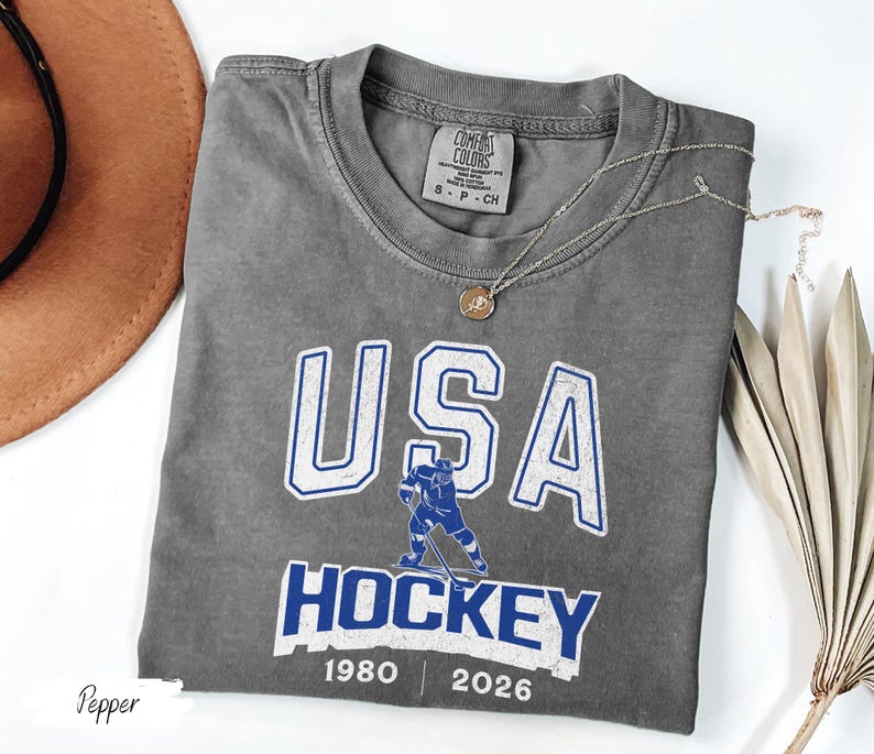 May include: A gray Comfort Colors t-shirt with a USA Hockey graphic in white and blue. The design includes a hockey player illustration and the years 1980 and 2026. A gold necklace is draped across the shirt.