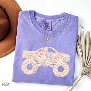 Op de afbeelding: Een violet Comfort Colors T-shirt met een bloemen monster truck ontwerp. De truck is gevuld met roze, witte en blauwe bloemen. Een gouden ketting en een bruine hoed zijn ook zichtbaar.