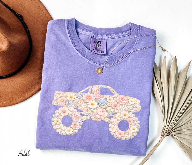 Op de afbeelding: Een violet Comfort Colors T-shirt met een bloemen monster truck ontwerp. De truck is gevuld met roze, witte en blauwe bloemen. Een gouden ketting en een bruine hoed zijn ook zichtbaar.