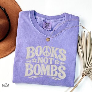 May include: A violet-colored t-shirt with the text "BOOKS NOT BOMBS" in a retro font. The design includes a peace sign and flowers. A gold necklace and a brown hat are also in the image.