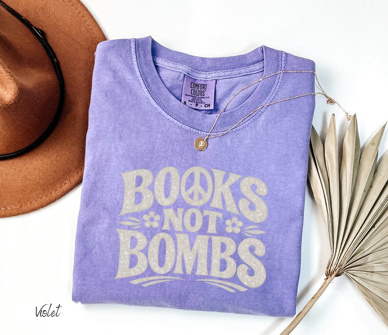 May include: A violet-colored t-shirt with the text "BOOKS NOT BOMBS" in a retro font. The design includes a peace sign and flowers. A gold necklace and a brown hat are also in the image.