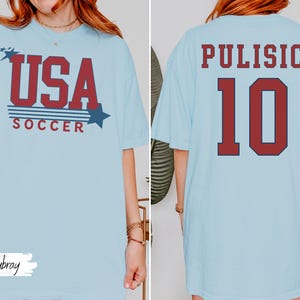 May include: Light blue t-shirt with "USA SOCCER" in red and blue text on the front. The back of the shirt displays "PULISIC 10" in red lettering. The shirt is a casual style.