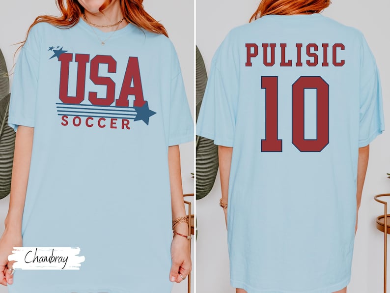 May include: Light blue t-shirt with "USA SOCCER" in red and blue text on the front. The back of the shirt displays "PULISIC 10" in red lettering. The shirt is a casual style.