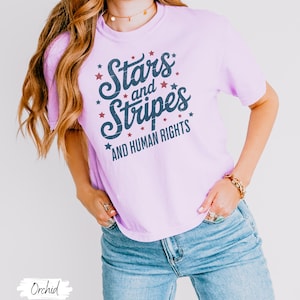 Puede incluir: Una camiseta de color lila claro con el texto "Stars and Stripes AND HUMAN RIGHTS" en escritura azul marino y estrellas rojas. La modelo lleva vaqueros azules. La camiseta es de estilo casual y c&oacute;modo.