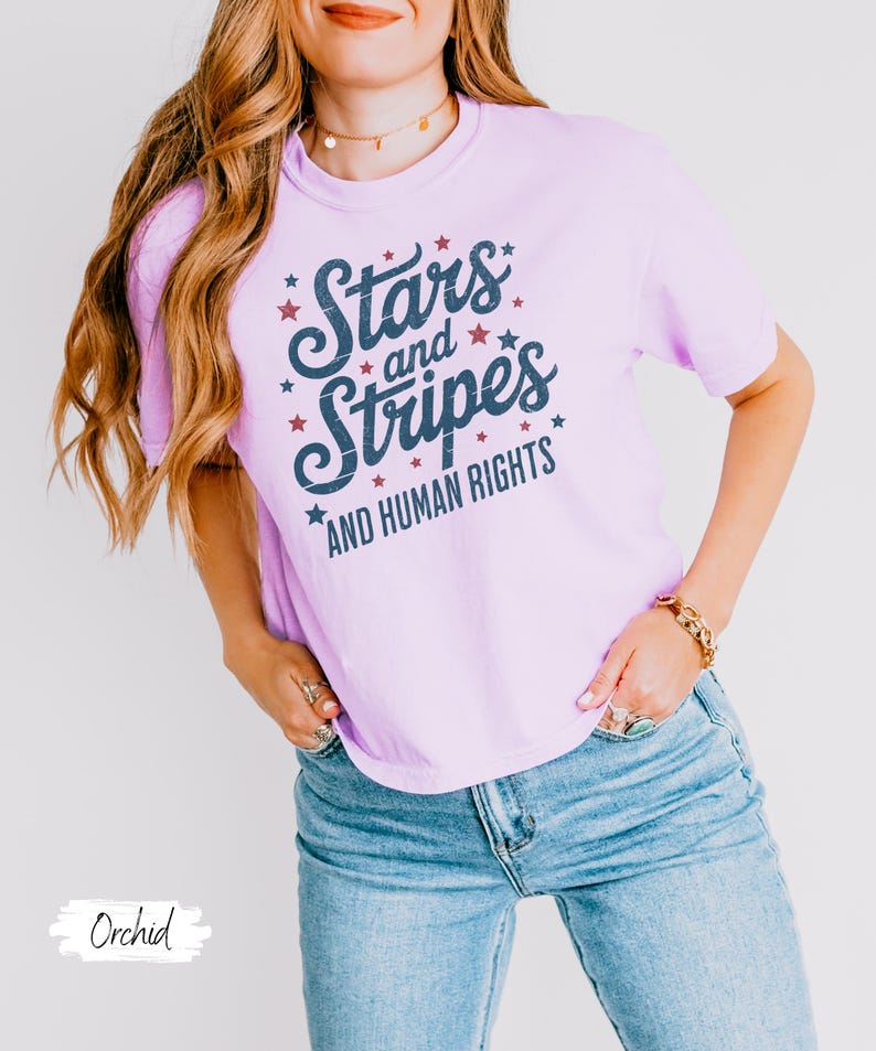 Puede incluir: Una camiseta de color lila claro con el texto "Stars and Stripes AND HUMAN RIGHTS" en escritura azul marino y estrellas rojas. La modelo lleva vaqueros azules. La camiseta es de estilo casual y c&oacute;modo.