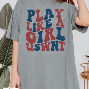 May include: A grey oversized t-shirt with the text "PLAY LIKE A GIRL USWNT" in red and blue letters.
