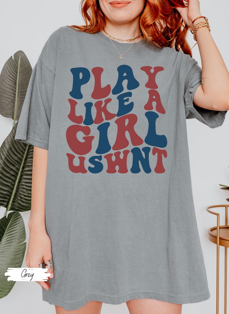 May include: A grey oversized t-shirt with the text "PLAY LIKE A GIRL USWNT" in red and blue letters.