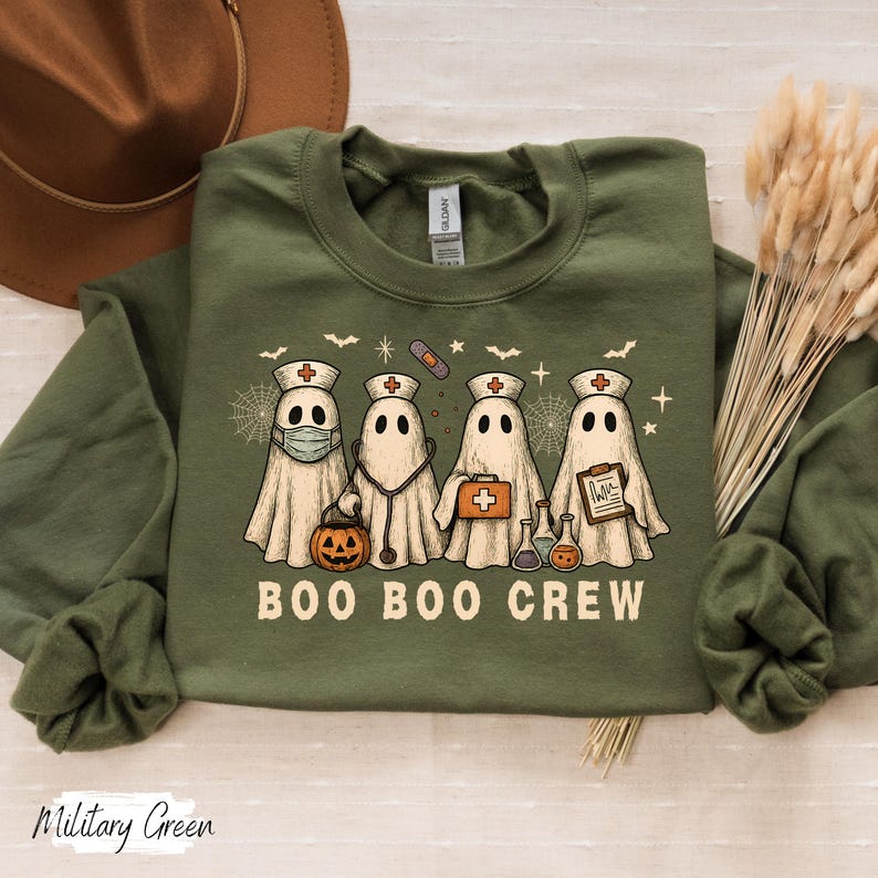 May include: Military green sweatshirt with a Halloween-themed design. The graphic features four ghost characters dressed as nurses, with the text "BOO BOO CREW" below. The ghosts are wearing medical accessories and holding a pumpkin.