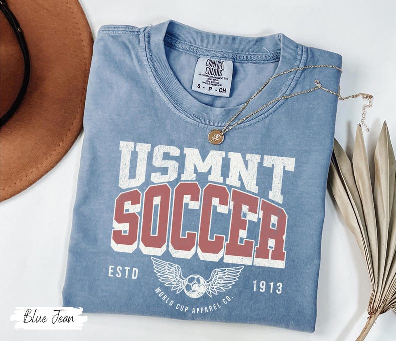 Może przedstawiać: Niebieska koszulka dżinsowa z napisem "USMNT SOCCER" w białych i czerwonych literach. Koszulka posiada piłkę nożną ze skrzydłami oraz napisy "ESTD 1913" i "WORLD CUP APPAREL CO." Złoty naszyjnik jest na koszulce.