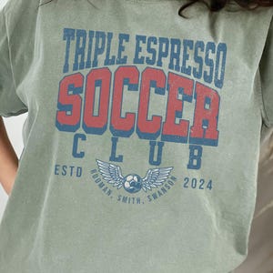 Op de afbeelding: Een groen T-shirt met de tekst "Triple Espresso Soccer Club" in rode en blauwe letters. De tekst "Estd 2024" staat onder de clubnaam. Een voetbal met vleugels bevindt zich in het midden van het shirt.