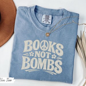 May include: A blue jean colored t-shirt with the text "BOOKS NOT BOMBS" in a retro font. The shirt has a crew neck and short sleeves. A gold necklace with a pendant is on the shirt. A brown hat is in the background.