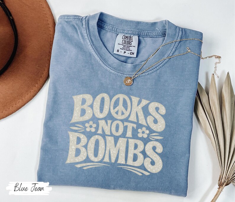 May include: A blue jean colored t-shirt with the text "BOOKS NOT BOMBS" in a retro font. The shirt has a crew neck and short sleeves. A gold necklace with a pendant is on the shirt. A brown hat is in the background.