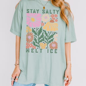 Op de afbeelding: Lichtgroen T-shirt met de tekst "STAY SALTY MELT ICE" in het groen. Het shirt heeft een bloemenontwerp met roze, gele, oranje en groene bloemen. Het T-shirt heeft korte mouwen en een losse pasvorm.