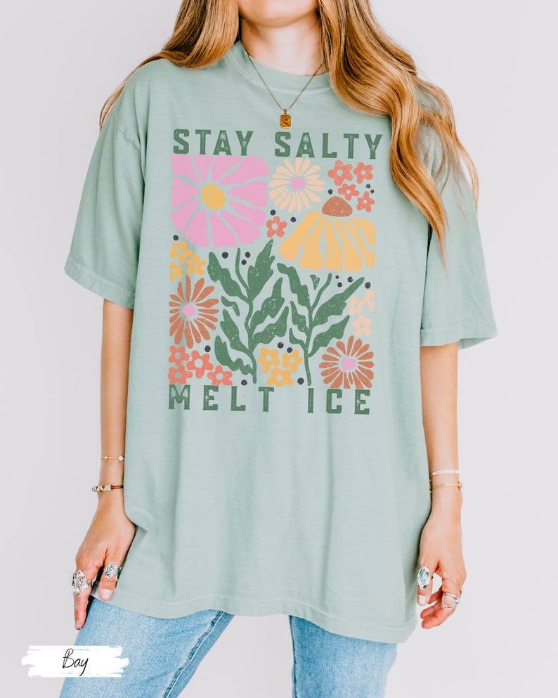 Op de afbeelding: Lichtgroen T-shirt met de tekst "STAY SALTY MELT ICE" in het groen. Het shirt heeft een bloemenontwerp met roze, gele, oranje en groene bloemen. Het T-shirt heeft korte mouwen en een losse pasvorm.