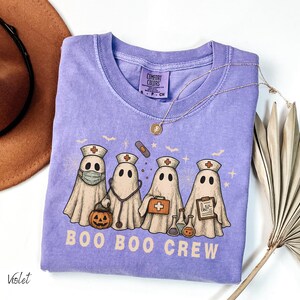 May include: A violet Comfort Colors t-shirt featuring a graphic of four ghost characters dressed as medical professionals. The ghosts are wearing nurse hats and holding medical tools. The text "BOO BOO CREW" is printed below the ghosts.