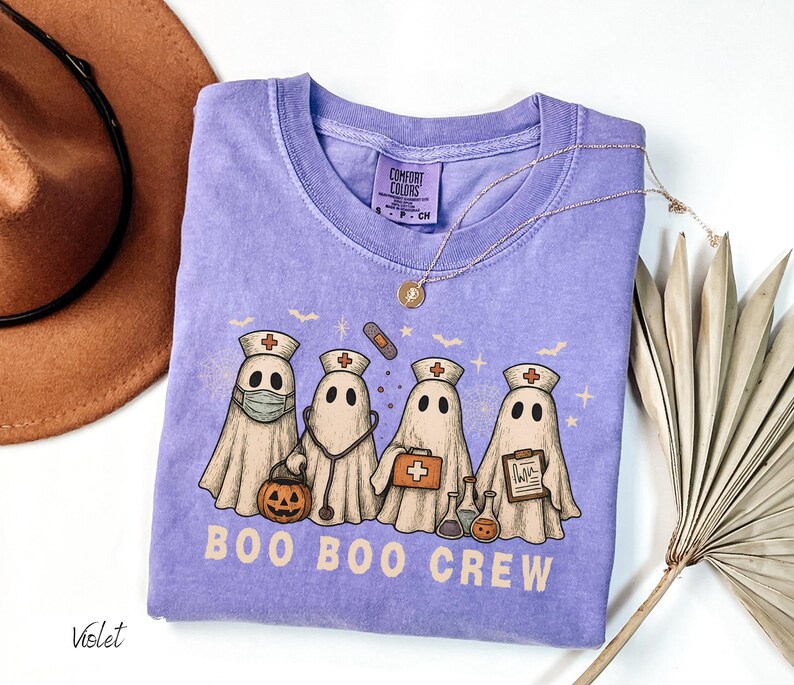 May include: A violet Comfort Colors t-shirt featuring a graphic of four ghost characters dressed as medical professionals. The ghosts are wearing nurse hats and holding medical tools. The text "BOO BOO CREW" is printed below the ghosts.