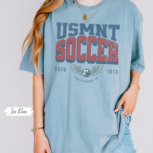 May include: Pale blue t-shirt featuring the text "USMNT SOCCER" in a retro design. The graphic includes a football with wings and the text "ESTD 1913 SPORTS APPAREL CO." The shirt is a light blue colour.
