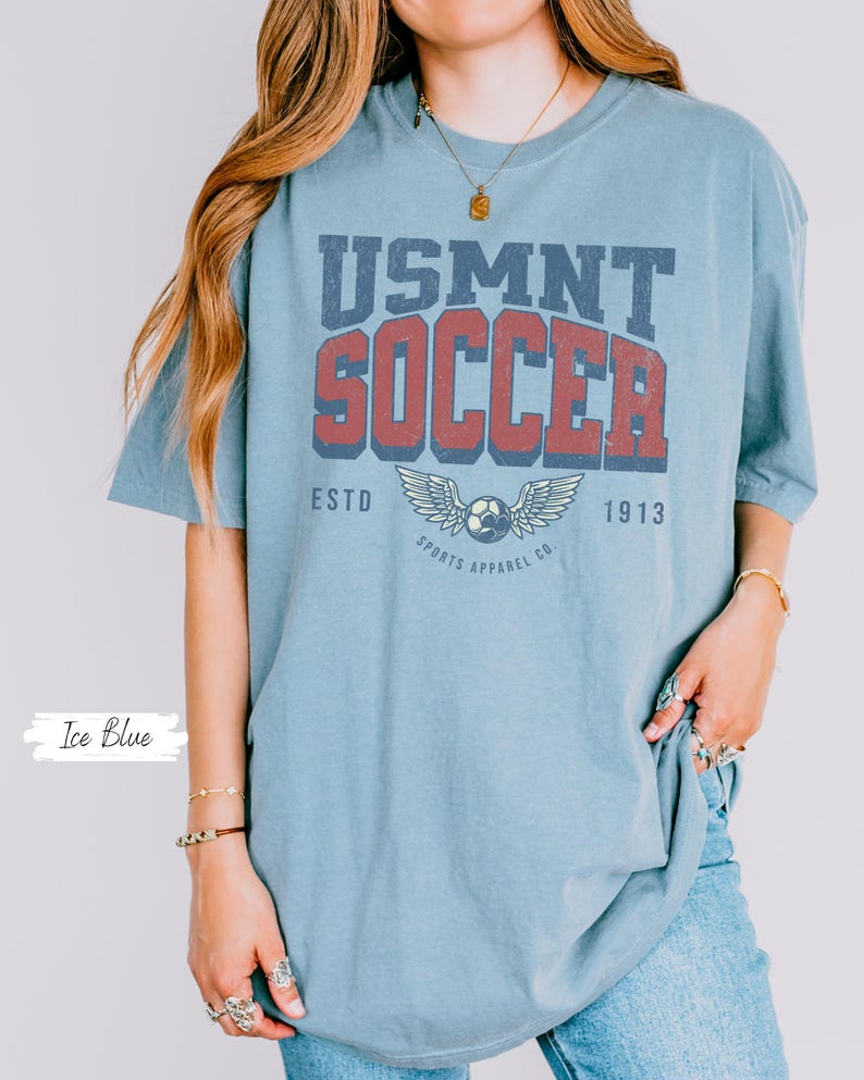 May include: Pale blue t-shirt featuring the text "USMNT SOCCER" in a retro design. The graphic includes a football with wings and the text "ESTD 1913 SPORTS APPAREL CO." The shirt is a light blue colour.