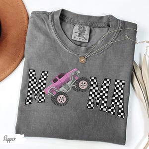 May include: A gray t-shirt with the word "MAMA" in a checkered pattern and a pink monster truck graphic. The shirt has a crew neck and short sleeves. A gold necklace is draped across the shirt.