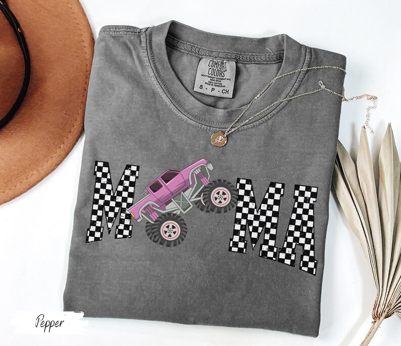 May include: A gray t-shirt with the word "MAMA" in a checkered pattern and a pink monster truck graphic. The shirt has a crew neck and short sleeves. A gold necklace is draped across the shirt.