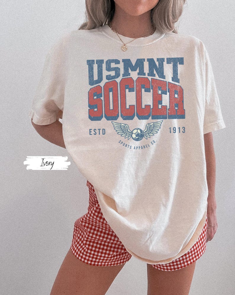May include: Cream-coloured t-shirt featuring the text "USMNT SOCCER" in blue and red, with a soccer ball graphic and wings. The shirt also has the text "ESTD 1913 SPORTS APPAREL CO." The model is wearing red and white checked shorts.