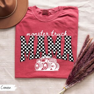 May include: Crimson-colored t-shirt with the words "monster truck MAMA" in a checkered pattern. A white graphic of a monster truck is below the text. The shirt is folded on a white surface.