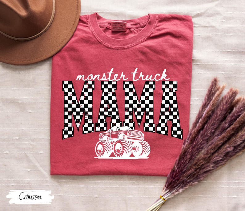 May include: Crimson-colored t-shirt with the words "monster truck MAMA" in a checkered pattern. A white graphic of a monster truck is below the text. The shirt is folded on a white surface.