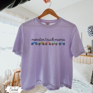May include: A light purple t-shirt with the text "monster truck mama" above a row of colorful monster truck illustrations. The shirt is hanging on a wooden hanger.