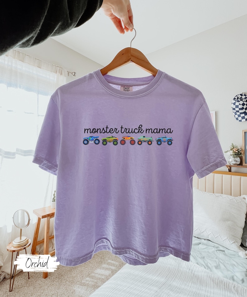 May include: A light purple t-shirt with the text "monster truck mama" above a row of colorful monster truck illustrations. The shirt is hanging on a wooden hanger.