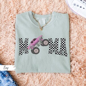 May include: A light green t-shirt with the word "MAMA" in black and white checkered letters. A pink monster truck graphic is centered between the letters. A gold necklace is visible at the neckline.