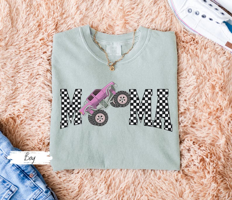 May include: A light green t-shirt with the word "MAMA" in black and white checkered letters. A pink monster truck graphic is centered between the letters. A gold necklace is visible at the neckline.